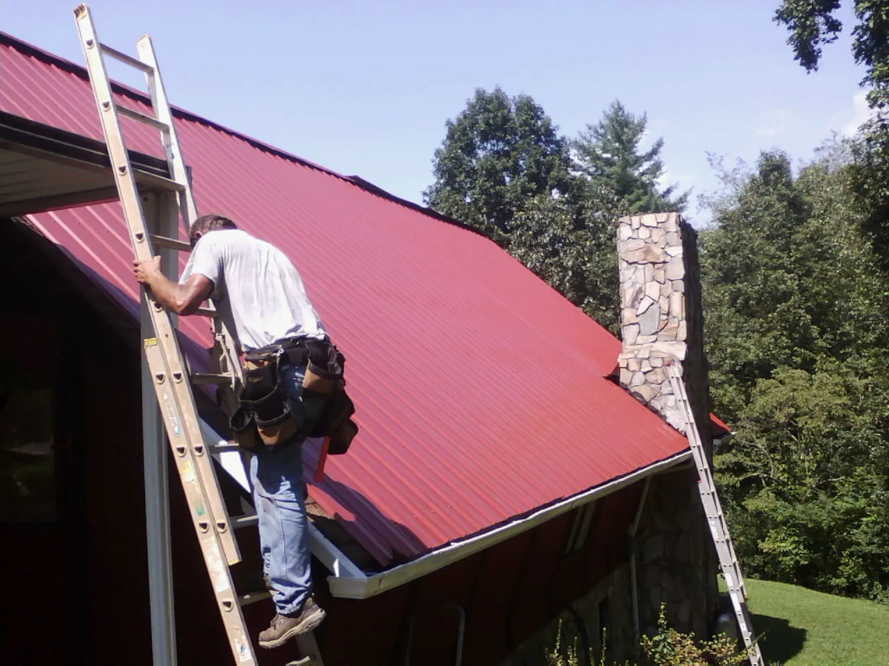 Roofer on a ladder beside a red metal roof and stone chimney — Soffit & Fascia Repair by Roofing Brothers in Barco
